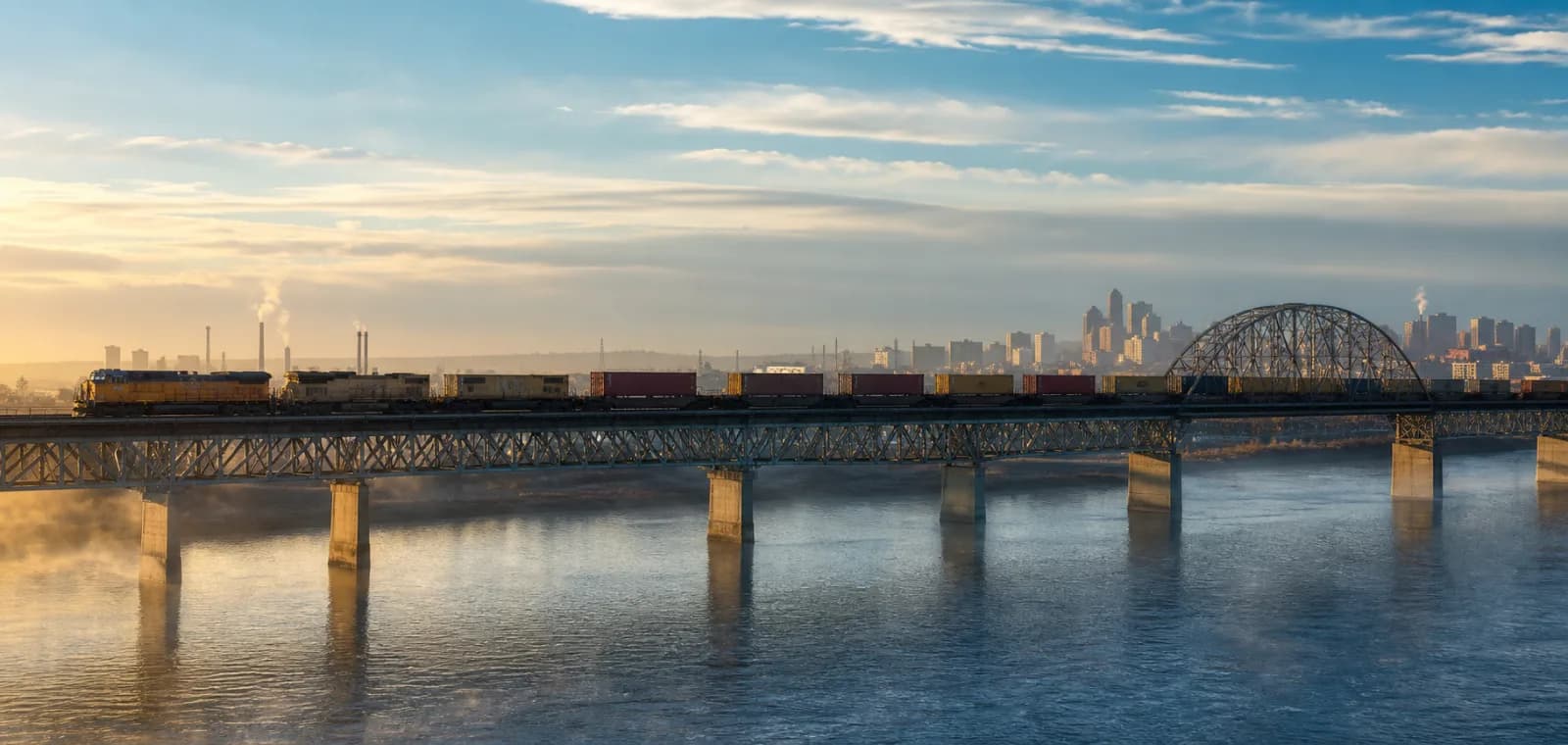 Freight train crossing a steel bridge between industrial border cities.