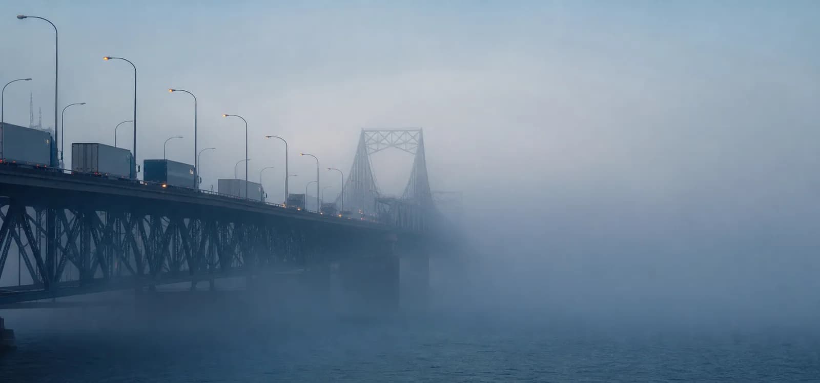 Trucks and bridge structures emerging through dense morning fog.